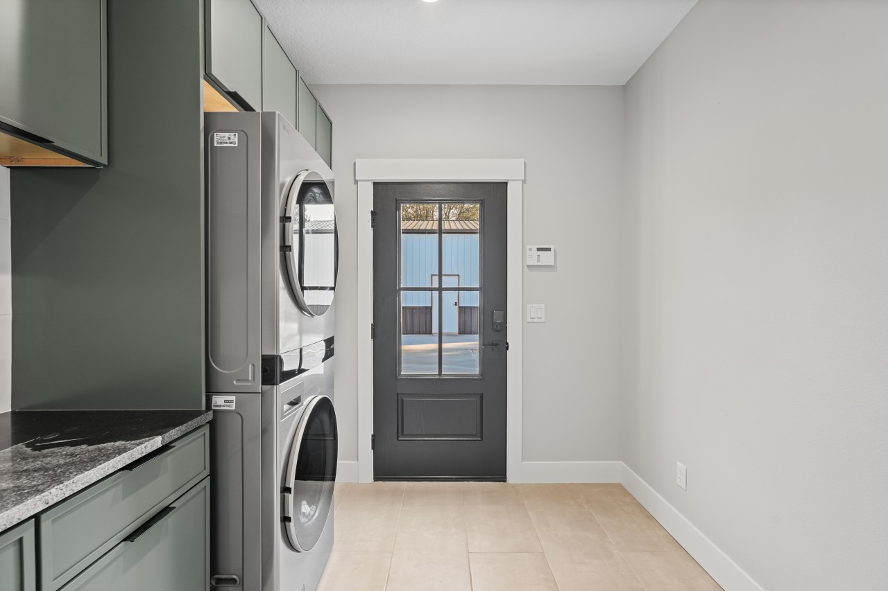 Custom laundry room with stacked front-load washer and dryer, dark green floor-to-ceiling cabinets and black glass entry door