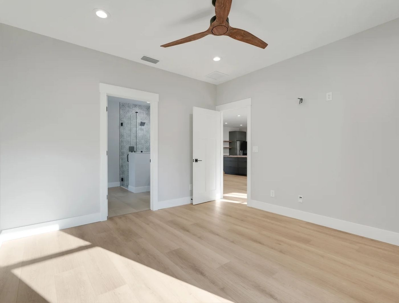 Bright master bedroom with light hardwood floors, ceiling fan and open doorways to en-suite bathroom and kitchen
