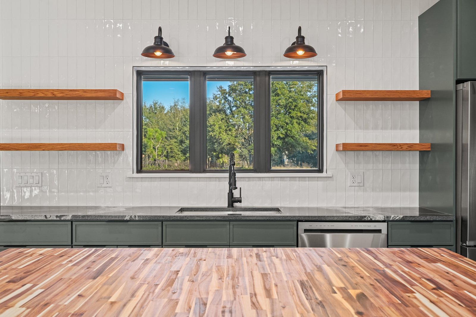 Kitchen sink area with black farmhouse faucet, sage green cabinets, butcher block island and white subway tile