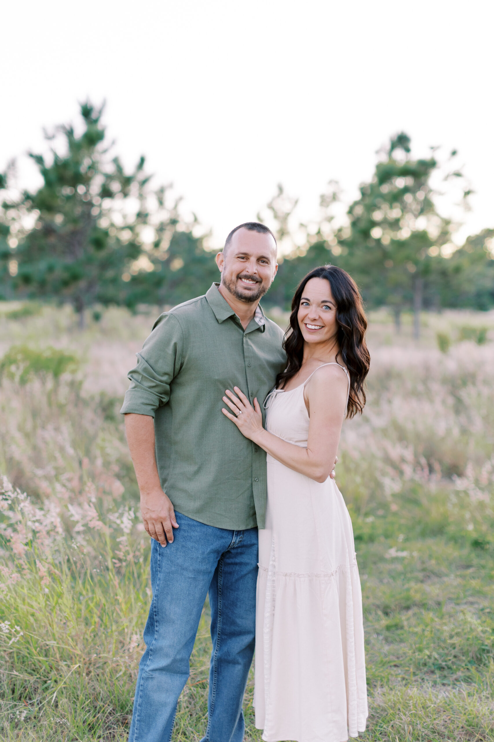 Smiling couple posing outdoors in golden-hour field, man in green shirt and jeans and woman in white sundress